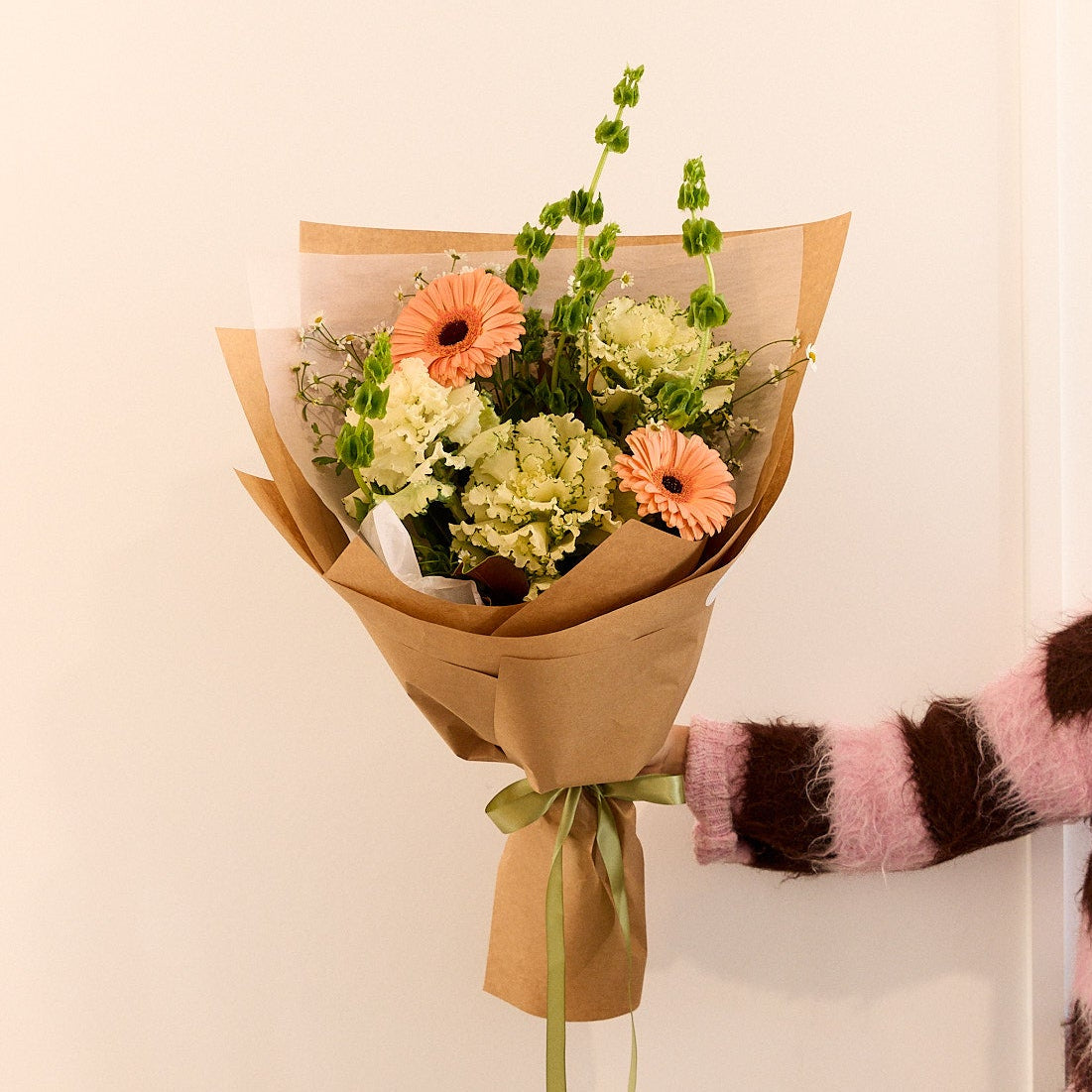A hand holding a small bouquet of flowers wrapped in brown kraft paper with a green ribbon, against a plain off-white wall. The person's arm is visible on the right, wearing a pink and brown striped fuzzy sweater.