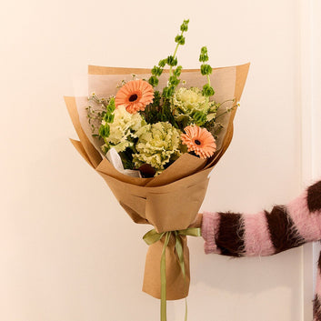 A hand holding a small bouquet of flowers wrapped in brown kraft paper with a green ribbon, against a plain off-white wall. The person's arm is visible on the right, wearing a pink and brown striped fuzzy sweater.