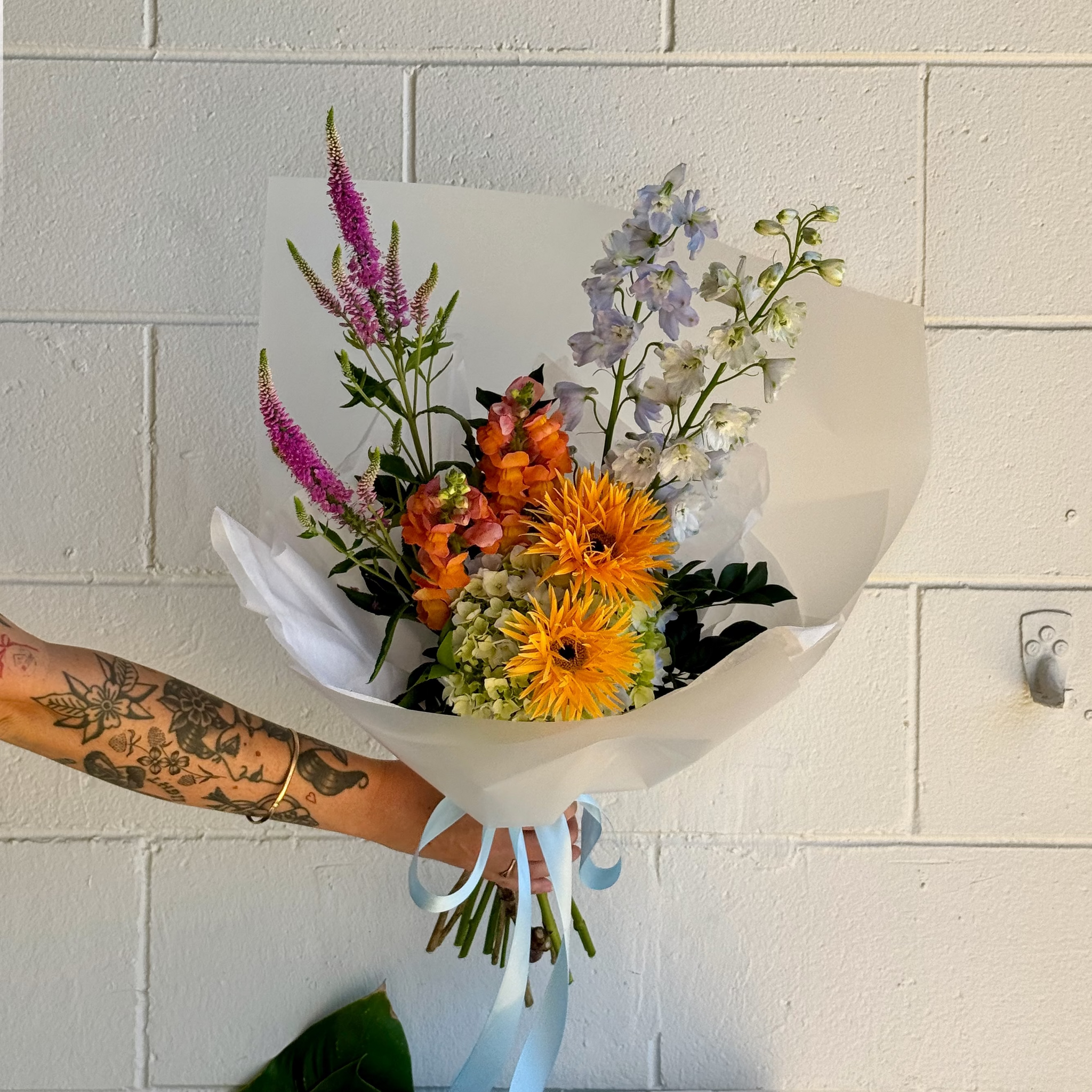 Bouquet of funky colourful flowers held by a person with tattoos against a tiled wall.