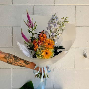 Bouquet of funky colourful flowers held by a person with tattoos against a tiled wall.