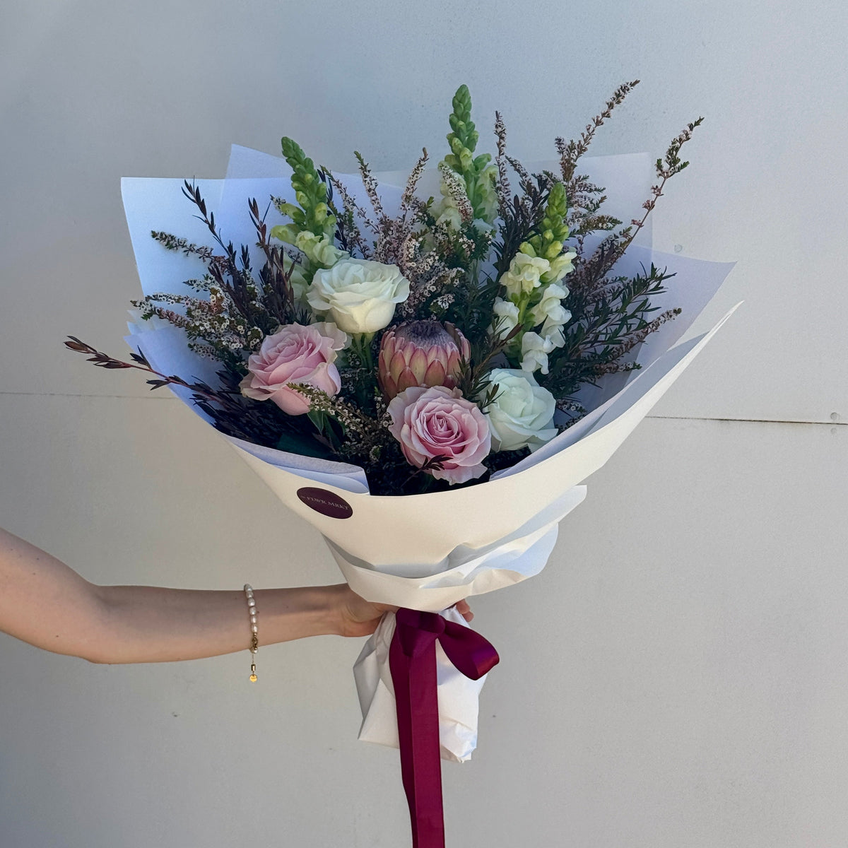 A hand holding a wrapped flower bouquet with pink roses, pale green blossoms, and green foliage, wrapped in white paper with a burgundy ribbon, against a plain light gray wall.