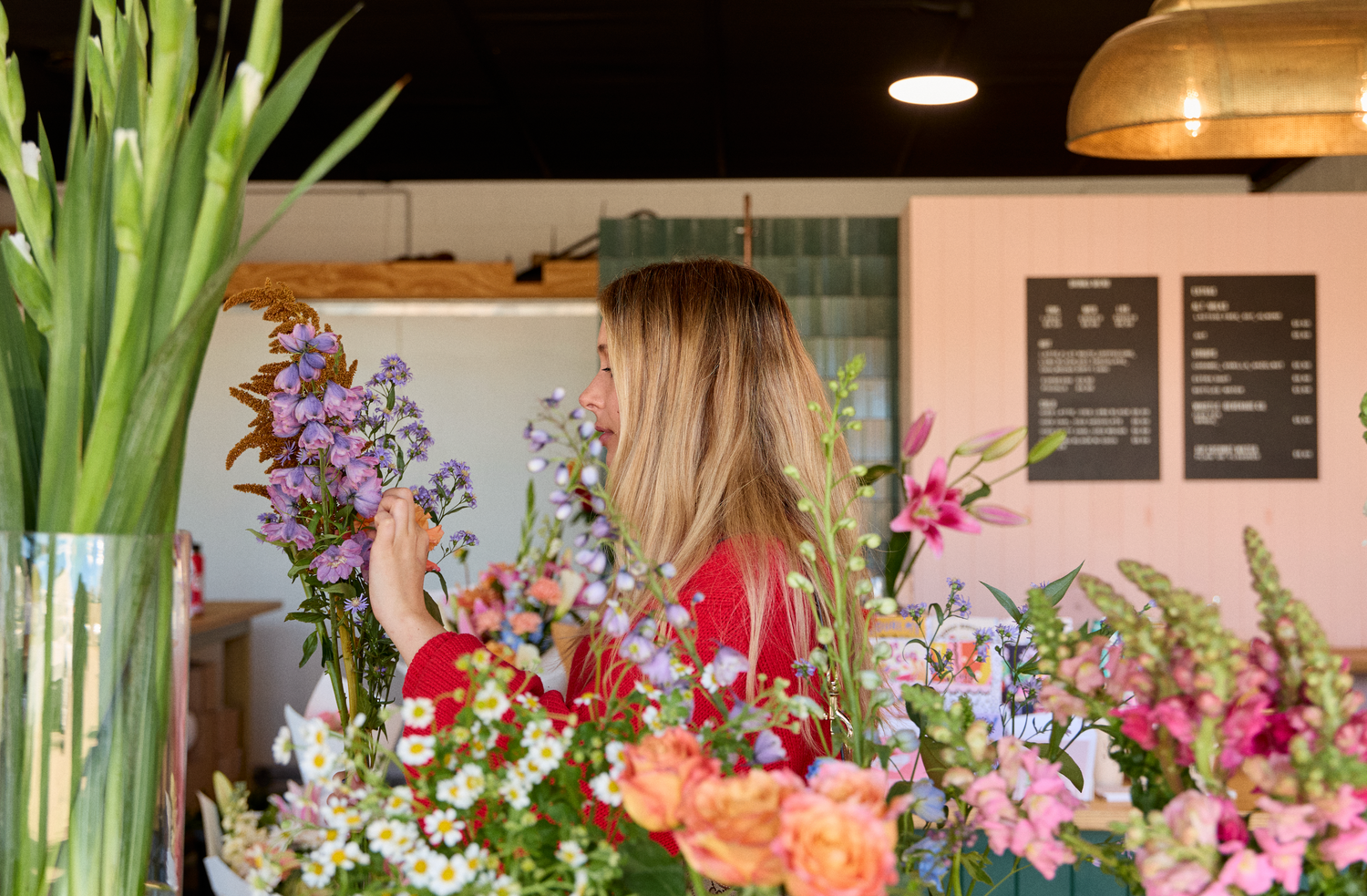 A woman in a red sweater stands in a flower shop surrounded by colorful flowers, holding a small bouquet of purple blossoms. The scene includes a large vase of green leaves on the left, a pink wall with black menu boards in the background, and warm hanging lights overhead.