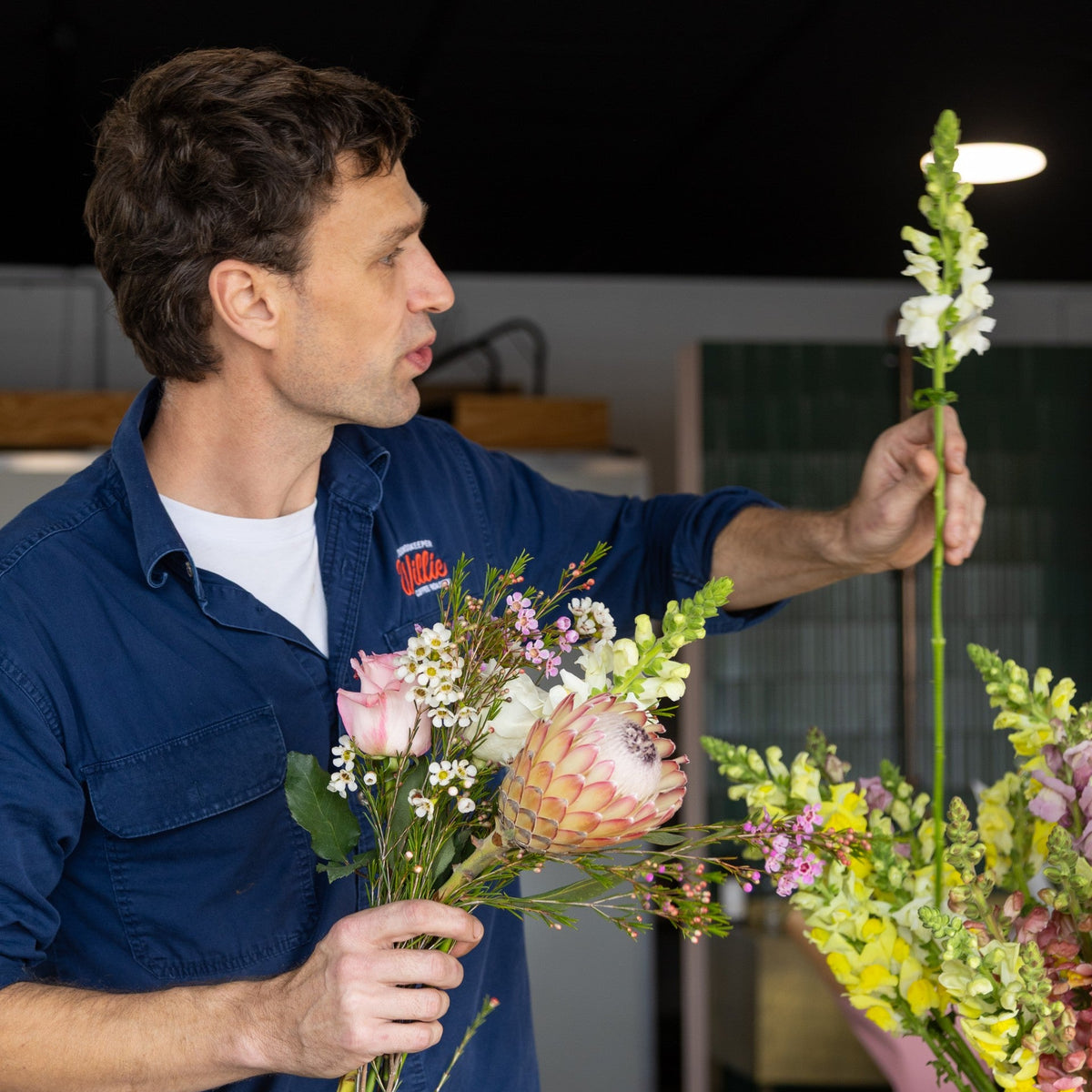 A man in a blue work shirt stands in a flower shop or studio, holding a bouquet of mixed flowers including a pink rose and a protea, while arranging a tall green stem in a vase of flowers on a table.